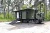 Militær lastebil med modulrom og rampe parkert på grusplass i skog