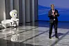 En robot og en mann står på en scene med blå bakgrunn og blankt gulv.