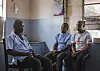 Tre menn sitter i et enkelt helserom med blå vegger og vindu. En T-skjorte med teksten «Malawi» er synlig.
