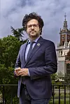 Mann i blå dress og briller står utendørs foran en historisk bygning med tårn.