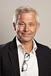 Mann i mørk blazer og hvit skjorte foran lys bakgrunn, med briller hengende i skjorten.