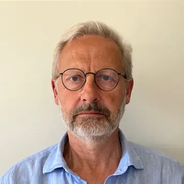 Mann med briller og grått skjegg foran lys vegg.