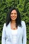 Kvinne i lys blazer og hvit topp står foran en grønn hekk.
