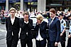 Fire personer i mørke klær står samlet foran en folkemengde utenfor en bygning.
