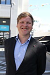 Smilende mann i mørk blazer står foran et skip ved en kai.
