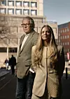Mann og kvinne i formelle klær står foran kontorbygninger i en by.