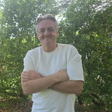 Smilende mann i hvit genser står med armene i kors foran busker.