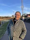 Kvinne med briller og mønstret kåpe står på en gangvei ved et anleggsområde.