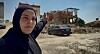 Kvinne i hijab peker mot ruiner og skadde bygninger i et urbant område.