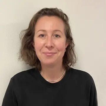 Kvinne med brunt, skulderlangt hår og svart genser foran en lys vegg.
