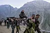 Fire menn bærer en pilegrim opp en fjellsti på en dandi, omgitt av høye fjell og andre vandrere.