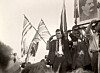 Menn med flagg og et stort portrett går fremst i en historisk folkemengde.