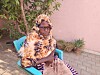 Kvinne med mønstret hodeplagg og solbriller sitter på en blå stol utendørs.