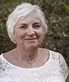 Eldre kvinne med kort hvitt hår og hvit blondetopp foran grønn vegetasjon.