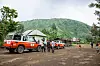 Oransje og hvite terrengkjøretøy står ved en bygning, med mennesker og en grønn fjellside i bakgrunnen.