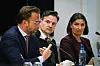 Tre personer sitter ved et bord med mikrofoner under en paneldebatt.