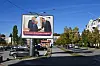 Billboard med to menn i dress ved en trafikkert bygate med biler og høsttrær.