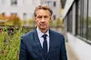 Mann i mørk dress står ved et moderne kontorbygg og blomstrende busker.